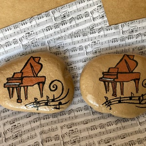 May include: Two painted rocks with a brown piano and musical notes on each. The rocks are on a sheet of music.