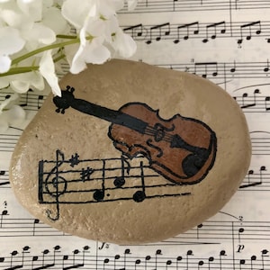 Puede incluir: Una piedra pintada con un viol&iacute;n marr&oacute;n y notas musicales sobre un fondo beige.