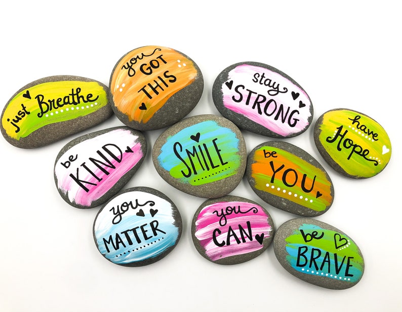 May include: Painted rocks with positive messages, including "you got this", "be kind", "smile", "stay strong", "have hope", "you matter", "you can", and "be brave".