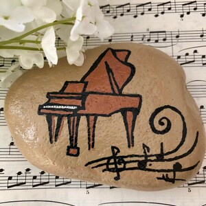 May include: A hand-painted rock with a brown grand piano and musical notes on a sheet of music.