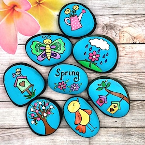 May include: Eight painted stones with spring themes. The stones are painted blue with various spring designs, including a butterfly, a birdhouse, a bird, a tree, a watering can, a flower, and a cloud with raindrops. The stones are arranged in a circle on a wooden surface.