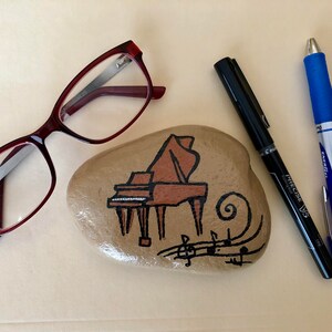 May include: A painted rock with a brown piano and musical notes. The rock is surrounded by a pair of eyeglasses, a black pen, and a blue pen.