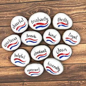 May include: A collection of painted rocks with positive words like "cheerful", "trustworthy", and "thrifty". Each rock is white with black dotted borders, red and blue wavy lines, and black lettering. The rocks are arranged on a wooden surface.
