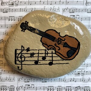 Puede incluir: Una piedra pintada a mano con un viol&iacute;n marr&oacute;n y notas musicales sobre un fondo beige.