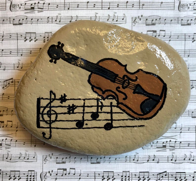 Puede incluir: Una piedra pintada a mano con un viol&iacute;n marr&oacute;n y notas musicales sobre un fondo beige.