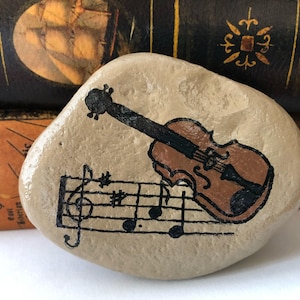 Puede incluir: Una piedra pintada a mano con un viol&iacute;n marr&oacute;n y notas musicales. La piedra es de color marr&oacute;n claro y tiene una textura rugosa.