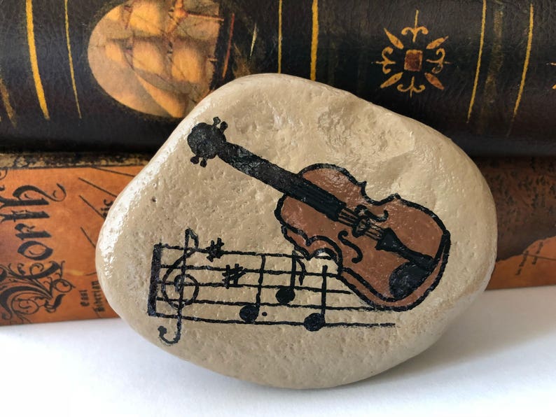 Puede incluir: Una piedra pintada a mano con un viol&iacute;n marr&oacute;n y notas musicales. La piedra es de color marr&oacute;n claro y tiene una textura rugosa.