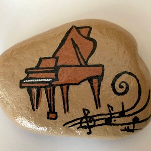 May include: A hand-painted rock with a brown grand piano and musical notes. The piano is painted in a cartoon style with black outlines and brown and white details. The musical notes are black and swirl around the piano.