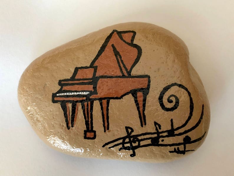 May include: A hand-painted rock with a brown grand piano and musical notes. The piano is painted in a cartoon style with black outlines and brown and white details. The musical notes are black and swirl around the piano.