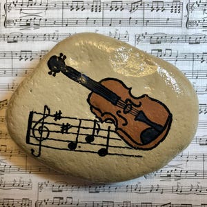 Puede incluir: Una piedra pintada con un viol&iacute;n marr&oacute;n y notas musicales sobre un fondo beige.