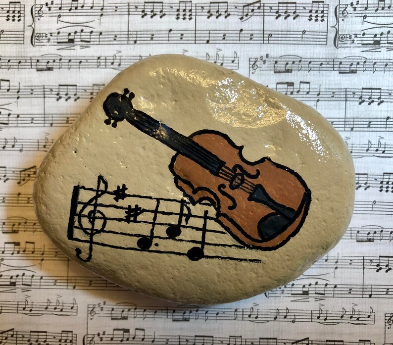 Puede incluir: Una piedra pintada con un viol&iacute;n marr&oacute;n y notas musicales sobre un fondo beige.