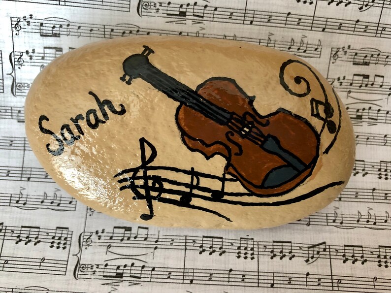 Puede incluir: Una piedra pintada con un viol&iacute;n marr&oacute;n, notas musicales y el nombre "Sarah" escrito en ella.