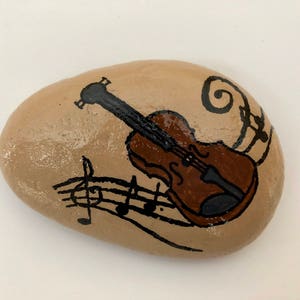 Puede incluir: Una piedra pintada con un viol&iacute;n marr&oacute;n y notas musicales sobre un fondo beige.