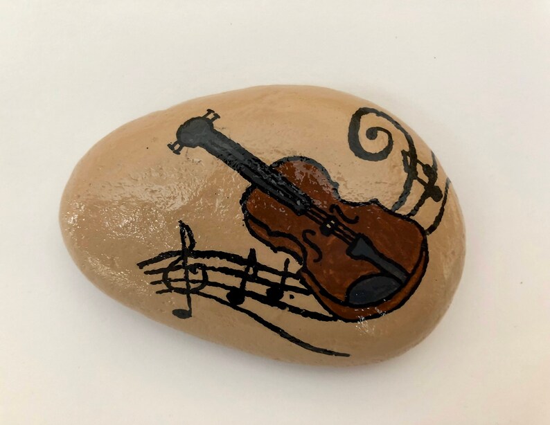 Puede incluir: Una piedra pintada con un viol&iacute;n marr&oacute;n y notas musicales sobre un fondo beige.