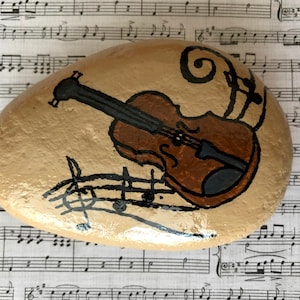 Puede incluir: Una piedra pintada a mano con un viol&iacute;n marr&oacute;n y notas musicales. La piedra est&aacute; sobre una hoja de m&uacute;sica.