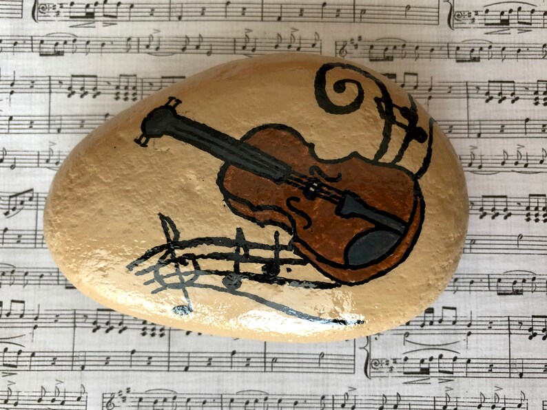 Puede incluir: Una piedra pintada a mano con un viol&iacute;n marr&oacute;n y notas musicales. La piedra est&aacute; sobre una hoja de m&uacute;sica.