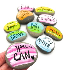May include: A set of painted rocks with inspirational messages. The rocks are painted in various colors, including pink, blue, green, yellow, and orange. The messages on the rocks include "You Can", "You Matter", "Be Brave", "Stay Strong", "Just Breathe", "Smile", "Be You", "Got This", "Have Hope", and "Kind".