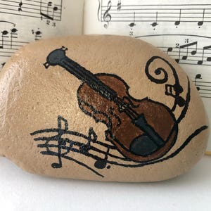 Puede incluir: Una piedra pintada con un viol&iacute;n marr&oacute;n y notas musicales sobre un fondo beige.