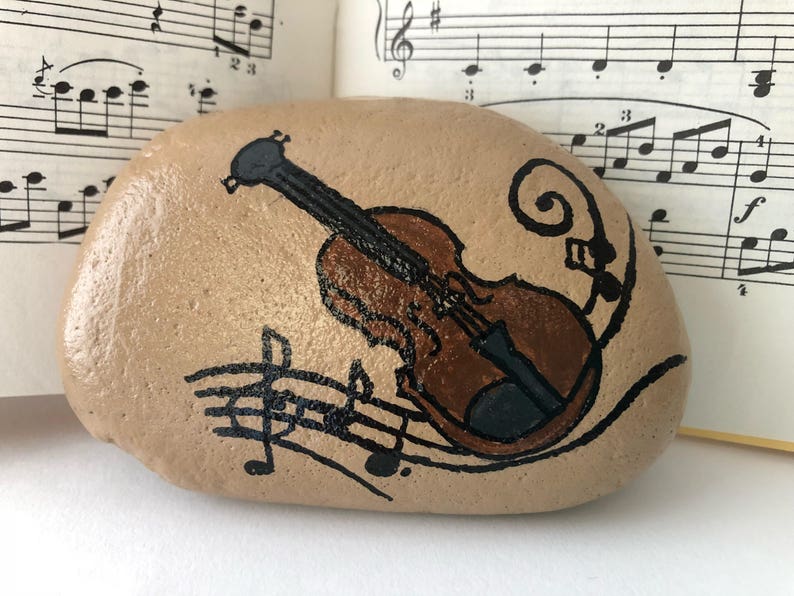 Puede incluir: Una piedra pintada con un viol&iacute;n marr&oacute;n y notas musicales sobre un fondo beige.