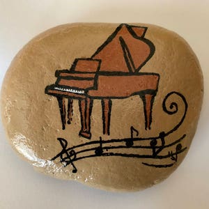May include: A hand-painted rock with a brown piano and musical notes painted on it. The piano is painted in a cartoon style with black outlines and brown and white details. The musical notes are painted in black.