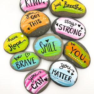May include: Ten painted rocks with inspirational messages, including "be kind", "you got this", "just breathe", "stay strong", "have hope", "be brave", "smile", "be you", "you can", and "you matter".