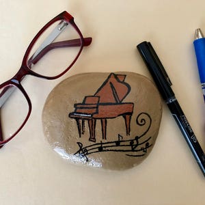 May include: A painted rock with a brown grand piano and musical notes. The rock is sitting on a beige surface with a pair of red glasses and two pens.