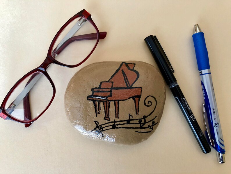 May include: A painted rock with a brown grand piano and musical notes. The rock is sitting on a beige surface with a pair of red glasses and two pens.