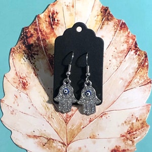 May include: Silver-coloured Hamsa hand earrings with clear crystal detailing and a blue eye. The earrings are presented on a black tag, set against a leaf-shaped paper cutout and a light blue background.