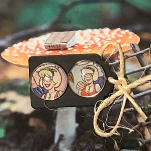 Puede incluir: Pendientes de botón redondos con un personaje de dibujos animados con un atuendo rojo y blanco. Los pendientes están colocados sobre un respaldo rectangular negro. El fondo incluye una seta y una pequeña estructura de madera.