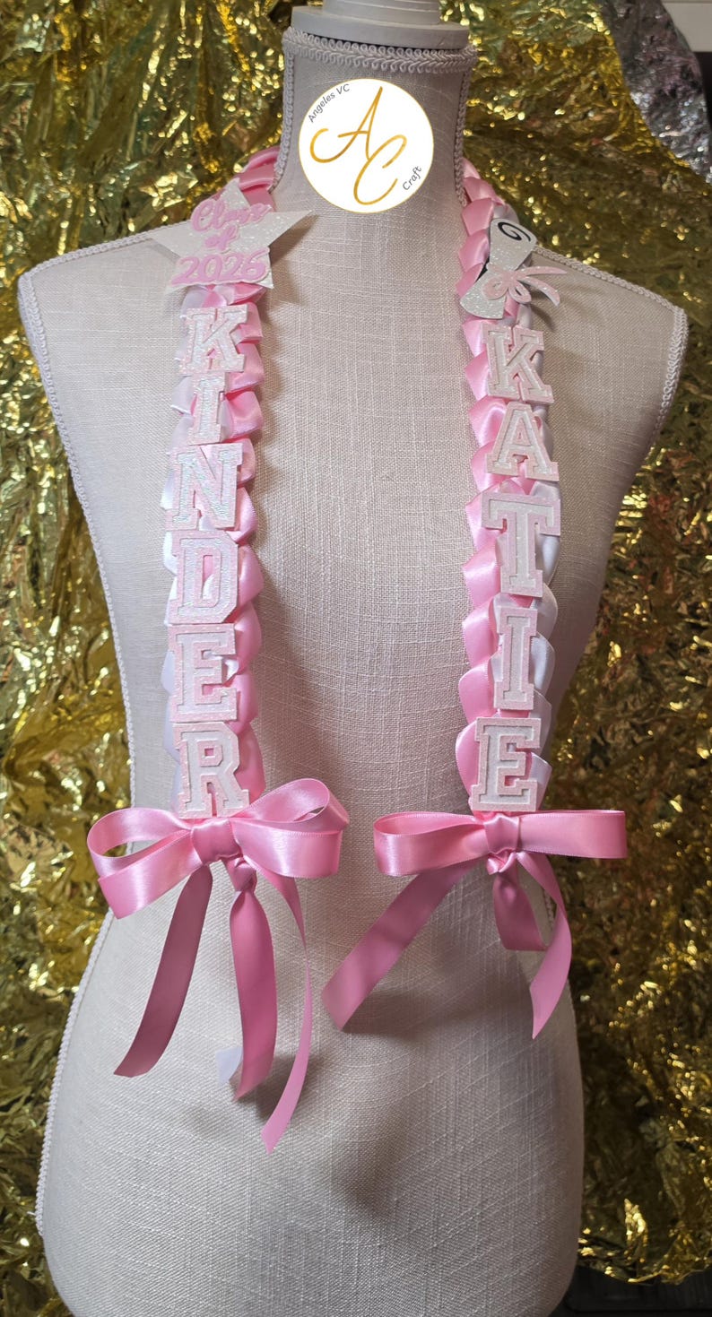 K&ouml;nnte beinhalten: Rosa Abschluss-Leis mit wei&szlig;en Blockbuchstaben, die "KINDER" und "KATIE" buchstabieren. Jede Lei hat eine rosa Satinbandschleife. Die Leis werden auf einer Schaufensterpuppe ausgestellt. Das Jahr "2025" ist oben auf einer Lei zu sehen.