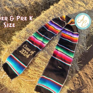May include: A black graduation stole with colorful stripes and the text "Class of 2024" in gold. The stole is designed for Kinder & Pre K students. The stole is lying on a hay bale.