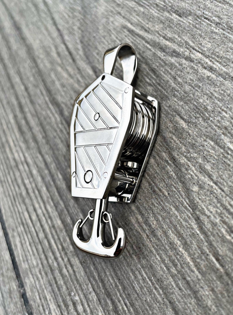Op de afbeelding: Een zilveren metalen sleutelhanger met een gedetailleerd ankerontwerp en een touwachtig wikkelmechanisme. De sleutelhanger is gevormd als een kist.
