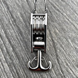Op de afbeelding: Een zilveren metalen sleutelhanger met een intrekbare haak en een veermechanisme. De haak heeft de vorm van een anker.
