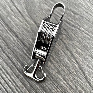 Op de afbeelding: Een zilveren metalen haak met een uniek ontwerp. De haak heeft een reeks ringen die kunnen worden gebruikt om een touw of ketting vast te maken. De haak is bevestigd aan een zilveren metalen anker.