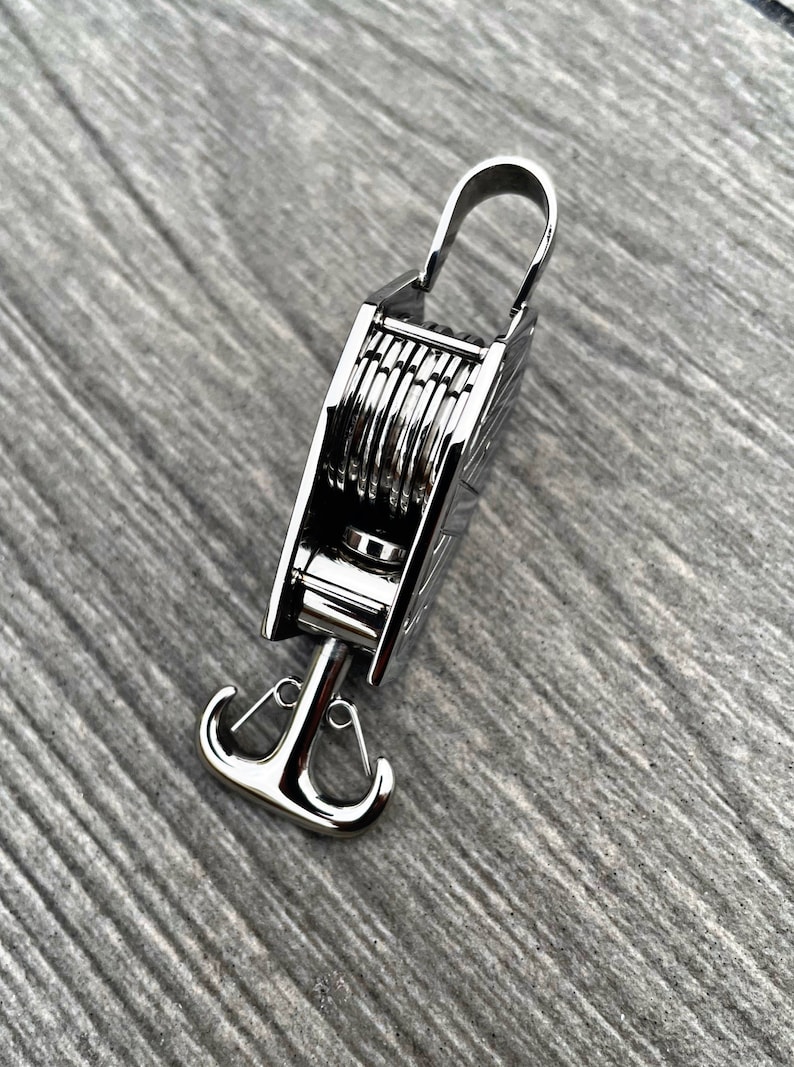 Op de afbeelding: Een zilveren metalen haak met een uniek ontwerp. De haak heeft een reeks ringen die kunnen worden gebruikt om een touw of ketting vast te maken. De haak is bevestigd aan een zilveren metalen anker.