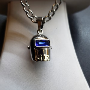 Può includere: Una collana d'argento con un ciondolo a forma di casco da saldatore in argento con una pietra preziosa blu e le iniziali "JR" in oro.