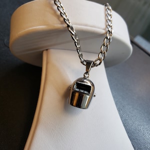 Peut inclure: Un collier en chaîne argent avec un pendentif en argent en forme de casque de soudeur.
