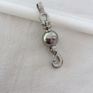 Op de afbeelding: Een zilveren metalen hanger met een ronde bal en een haak. De hanger heeft een decoratief ontwerp met een kleine lus aan de bovenkant.