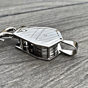 Op de afbeelding: Een zilveren metalen katrol met een haak en een lus. De katrol heeft een gegroefd wiel en een kooi eromheen.