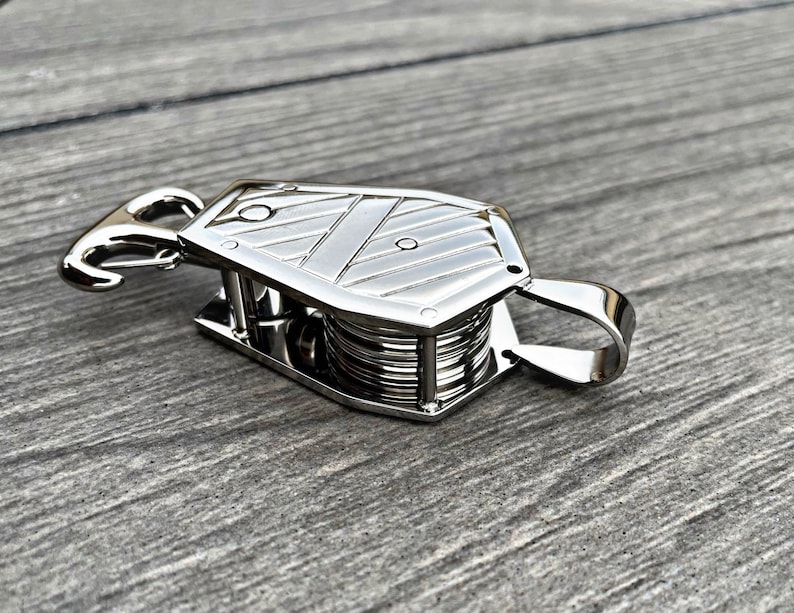 Op de afbeelding: Een zilveren metalen katrol met een haak en een lus. De katrol heeft een gegroefd wiel en een kooi eromheen.