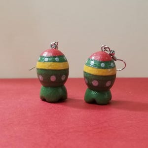 May include: Two handmade, colorful earrings. Each earring features a red top, a green band with white dots, a yellow band, a brown section with pink dots, and a green base. The earrings hang from silver-colored hooks.