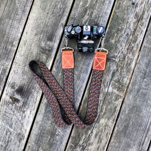 Pode incluir: Uma câmera preta com uma alça de couro marrom. A alça tem um padrão tecido colorido com fios vermelhos, amarelos e azuis.