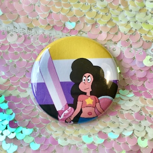 May include: A round button with a cartoon character holding a sword. The character has pink skin, brown hair, and is wearing a yellow, purple, and white shirt. The background of the button is a yellow, purple, and white striped pattern.