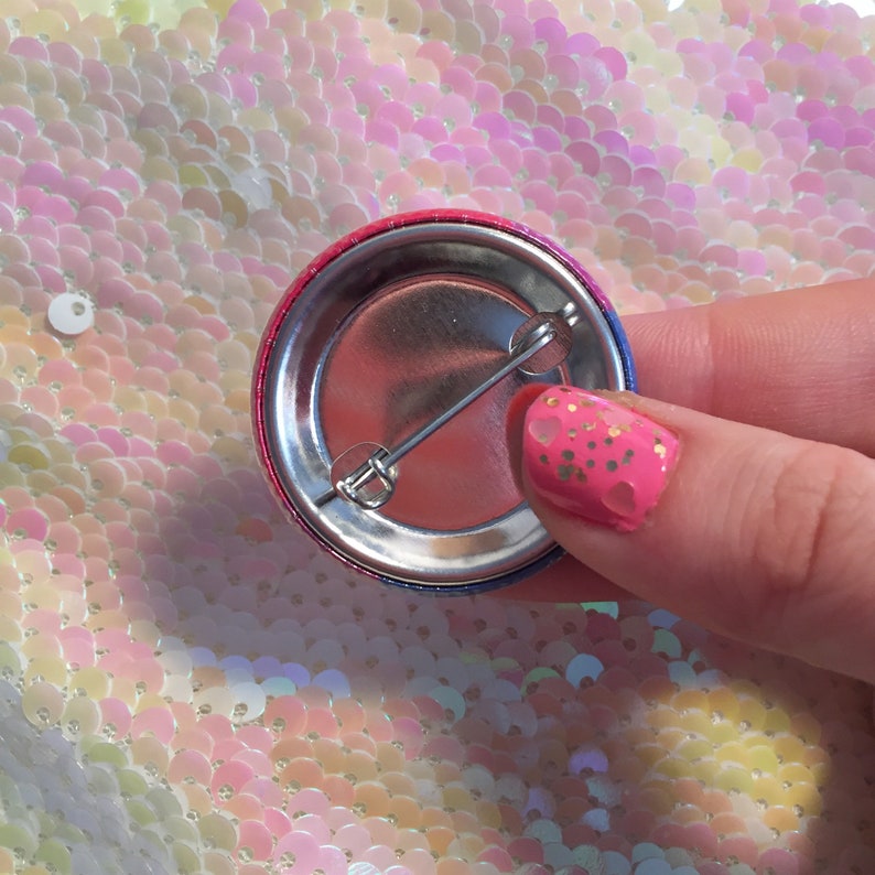 Puede incluir: Un bot&oacute;n redondo con un dise&ntilde;o degradado en rojo, rosa y azul. El pin tiene una parte trasera de metal con un cierre de imperdible. El bot&oacute;n est&aacute; sostenido por una mano con esmalte de u&ntilde;as rosa.