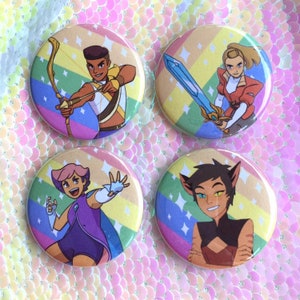 May include: Four round button badges featuring characters from the animated series She-Ra and the Princesses of Power. Each badge features a different character against a rainbow background with glitter accents. The characters are Adora, Catra, Glimmer, and Bow.