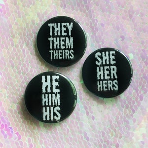 Puede incluir: Tres botones negros con texto blanco. Los botones dicen "They Them Theirs", "She Her Hers" y "He Him His".
