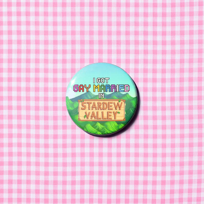 Puede incluir: Un bot&oacute;n redondo con el texto "I GOT GAY MARRIED IN STARDREW VALLEY" en fuente pixelada. El bot&oacute;n tiene un fondo de cielo azul claro con colinas verdes y un letrero de madera. El fondo es un patr&oacute;n de cuadros rosas y blancos.