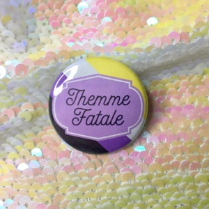 Könnte beinhalten: Ein lila, gelber, weißer und schwarzer Knopf mit dem Text "Themme Fatale" in einem weißen Rahmen.