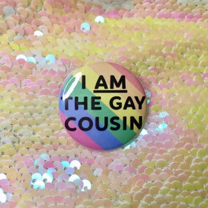 Puede incluir: Un botón de colores arcoíris con el texto "I AM THE GAY COUSIN" en negro.