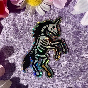 May include: A holographic sticker of a black and white skeleton unicorn with a rainbow glitter finish.